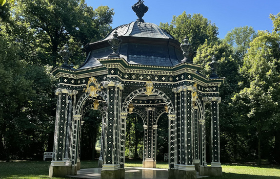 Ein gemauerter Pavillon in einem grünen Park, dunkelgrün gestrichen mit geometrischen Mustern in Gold. 